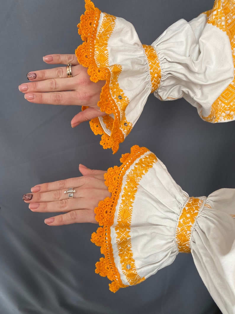 May include: White blouse with long sleeves and yellow embroidered details. The sleeves have a ruffled design and a crocheted trim.