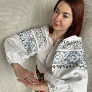 May include: A white embroidered blouse with a colorful woven belt. The blouse has a traditional Ukrainian design with blue and green floral patterns.