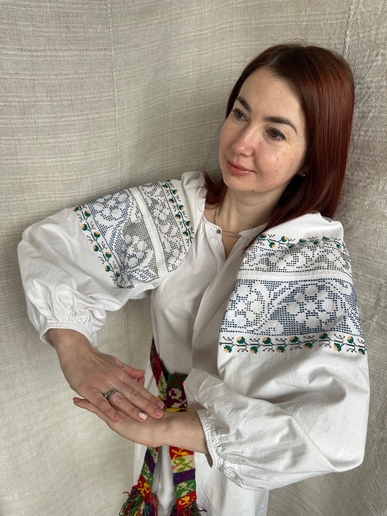May include: A white embroidered blouse with a colorful woven belt. The blouse has a traditional Ukrainian design with blue and green floral patterns.