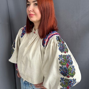 May include: A white linen blouse with embroidered sleeves. The embroidery is in shades of blue, green, red, and black. The blouse has a round neckline and long sleeves.
