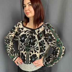 May include: A white, long-sleeved blouse with a black and green embroidered floral pattern. The blouse has a gathered neckline and a loose fit.