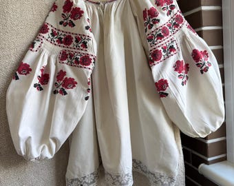 May include: An off-white blouse featuring voluminous long sleeves with intricate red and black floral embroidery. The garment has a gathered neckline with a thin red trim and a wide band of white lace detailing along the bottom hemline.
