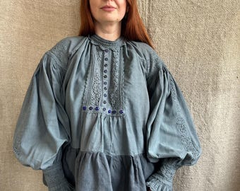 May include: A blue-grey blouse with intricate embroidery and puffed sleeves. The blouse features a high collar, a row of buttons down the front, and delicate lace detailing. The sleeves are gathered at the wrists, and the fabric has a slightly textured appearance.