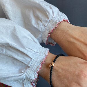 May include: A white shirt with red stitching around the cuffs. A black leather bracelet with a gold charm is worn on the wrist.