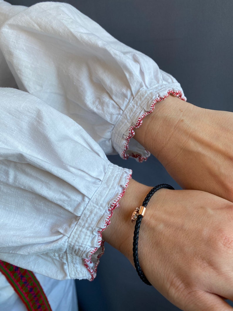 May include: A white shirt with red stitching around the cuffs. A black leather bracelet with a gold charm is worn on the wrist.