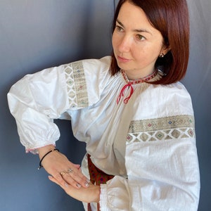 May include: A white embroidered blouse with long sleeves and a red ribbon at the neck. The blouse has a traditional folk design with geometric patterns in beige and brown.