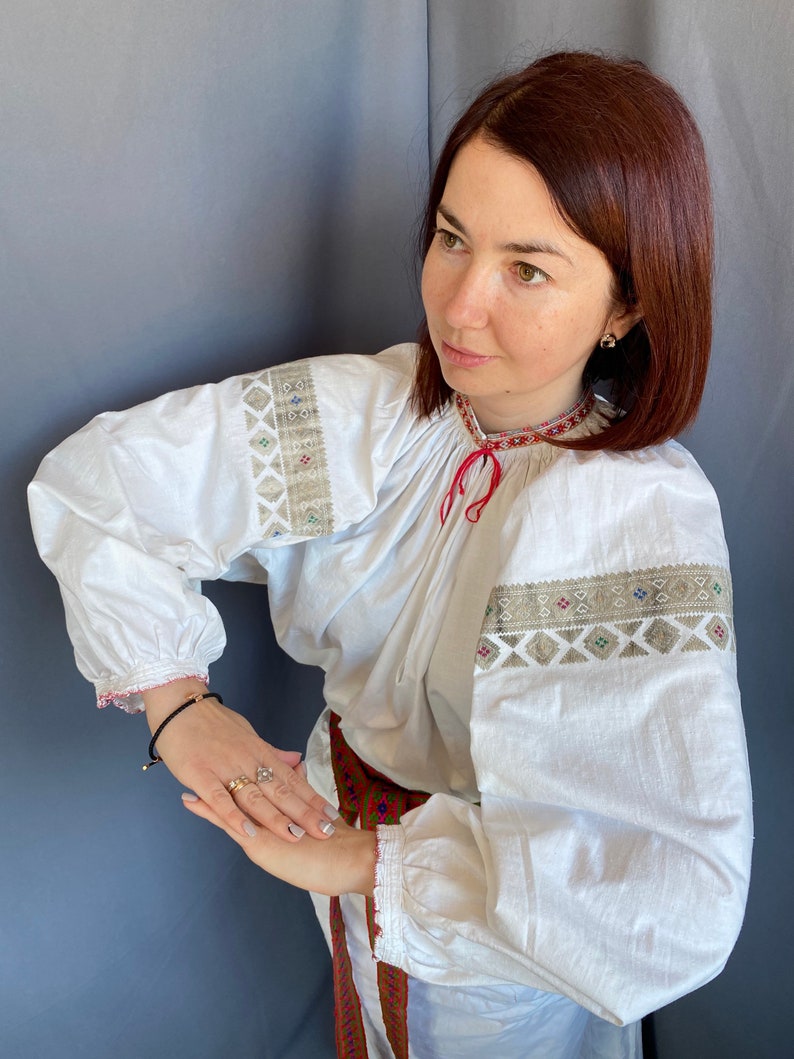 May include: A white embroidered blouse with long sleeves and a red ribbon at the neck. The blouse has a traditional folk design with geometric patterns in beige and brown.