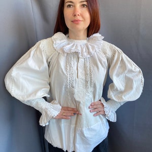 K&ouml;nnte beinhalten: Eine wei&szlig;e Baumwollbluse mit einem gro&szlig;en, gekr&auml;uselten Kragen und langen &Auml;rmeln. Die Bluse hat zarte Spitzendetails am Kragen und an den &Auml;rmeln.