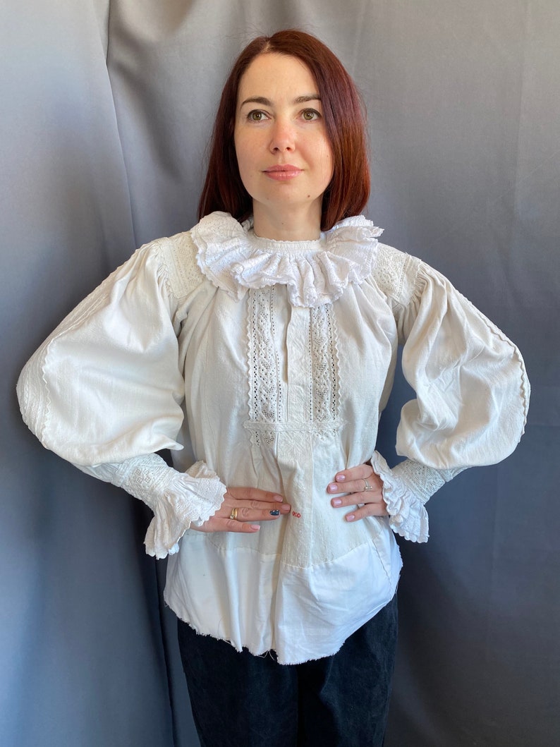 K&ouml;nnte beinhalten: Eine wei&szlig;e Baumwollbluse mit einem gro&szlig;en, gekr&auml;uselten Kragen und langen &Auml;rmeln. Die Bluse hat zarte Spitzendetails am Kragen und an den &Auml;rmeln.
