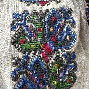 May include: Close-up of a white fabric with a detailed embroidered design. The design features a floral motif with blue, green, red, and yellow thread.