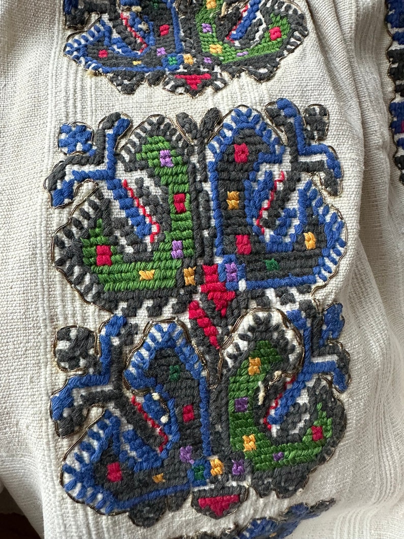 May include: Close-up of a white fabric with a detailed embroidered design. The design features a floral motif with blue, green, red, and yellow thread.