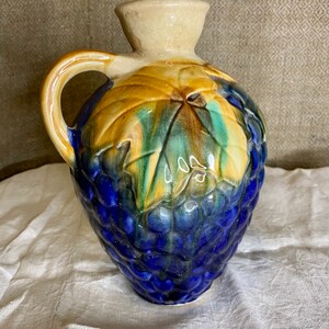 May include: A blue and brown ceramic pitcher with a grape design. The pitcher has a handle and a wide mouth.