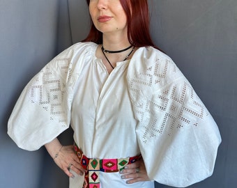 May include: A white embroidered blouse with long, puffy sleeves and a colorful woven belt. The blouse has a round neckline and a relaxed fit.