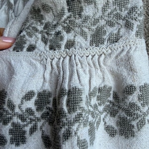 May include: A light beige linen-textured garment featuring dark olive green cross-stitch floral embroidery. The fabric displays a visible weave, with gathered pleats along a horizontal seam accented by a decorative zigzag stitch detail.