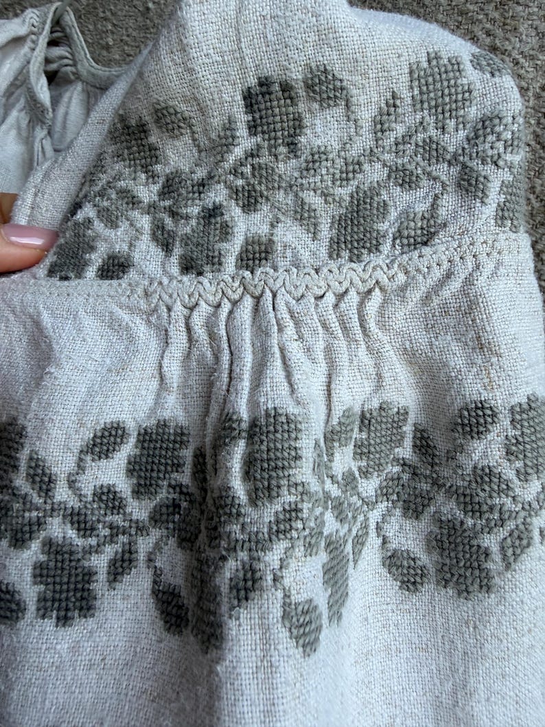 May include: A light beige linen-textured garment featuring dark olive green cross-stitch floral embroidery. The fabric displays a visible weave, with gathered pleats along a horizontal seam accented by a decorative zigzag stitch detail.