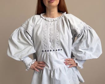 May include: A light blue long-sleeved blouse featuring voluminous puffed sleeves, a ruffled collar, and intricate white eyelet lace detailing down the center front. The garment includes a row of small dark blue buttons and ruffled cuffs.