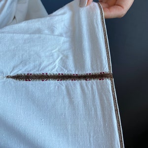 May include: Close-up of a white garment with a brown and red embroidered trim. The trim is a single line of stitching with a brown thread and a red thread running through it.