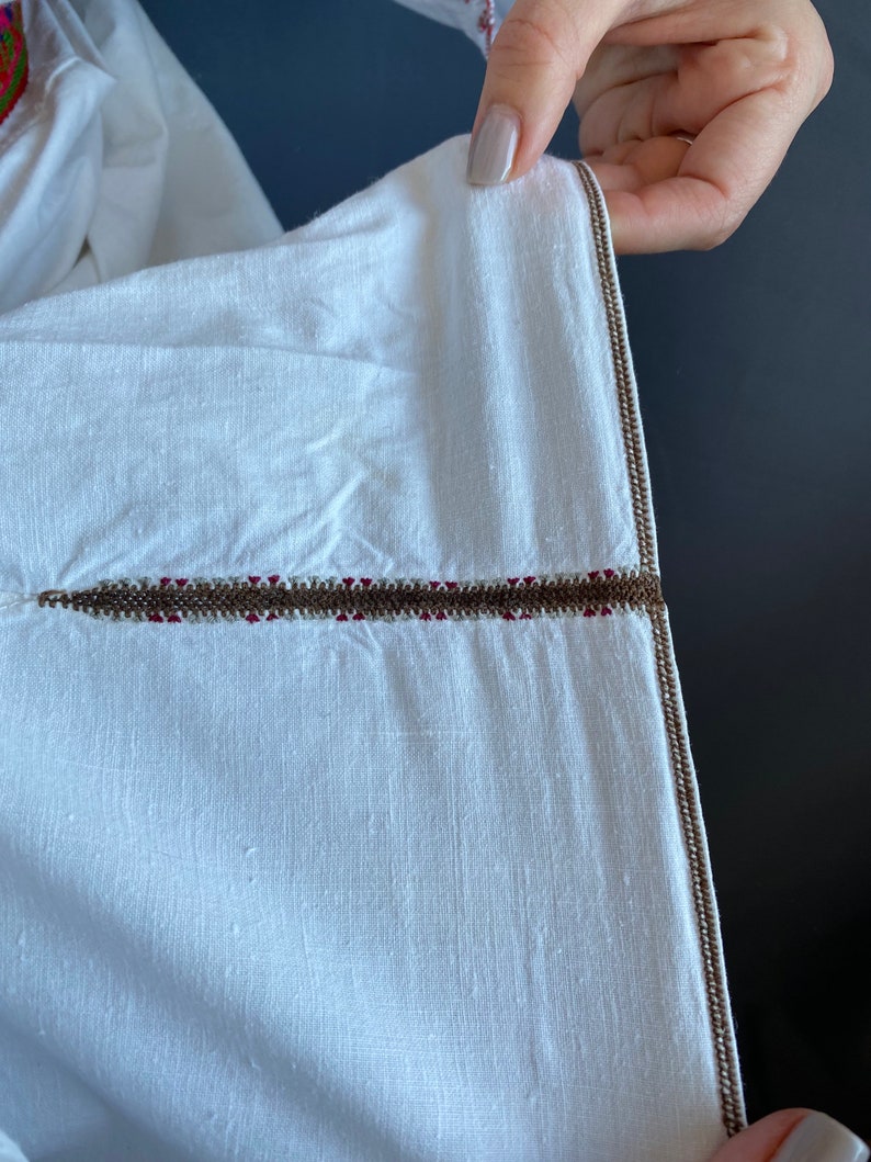 May include: Close-up of a white garment with a brown and red embroidered trim. The trim is a single line of stitching with a brown thread and a red thread running through it.