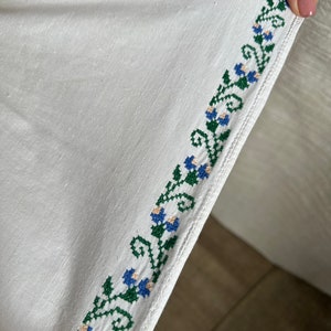 May include: White linen fabric with a green and blue embroidered floral border. The embroidery is a repeating pattern of stylized flowers and leaves.