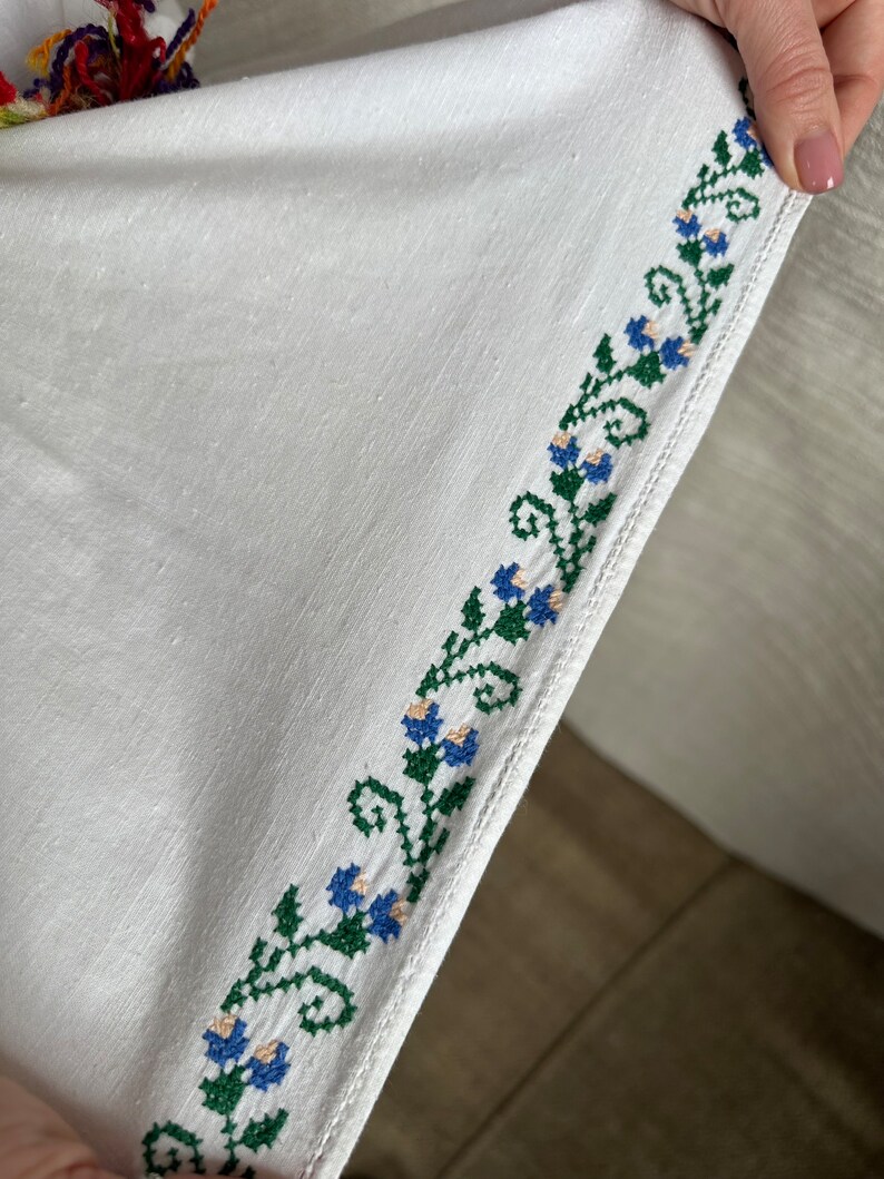 May include: White linen fabric with a green and blue embroidered floral border. The embroidery is a repeating pattern of stylized flowers and leaves.