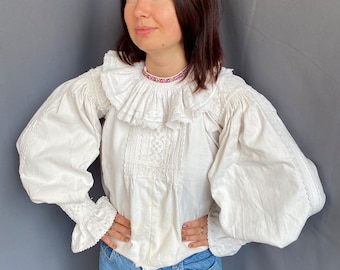 May include: A white blouse with a large, ruffled collar and long, puffed sleeves. The blouse is made of a delicate fabric and has intricate embroidery details. The model is wearing blue jeans.