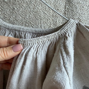 May include: A light beige linen blouse featuring a gathered neckline with intricate crochet detailing along the collar edge. The garment has long, voluminous sleeves and a textured fabric weave, displayed on a white wire hanger against a neutral background.