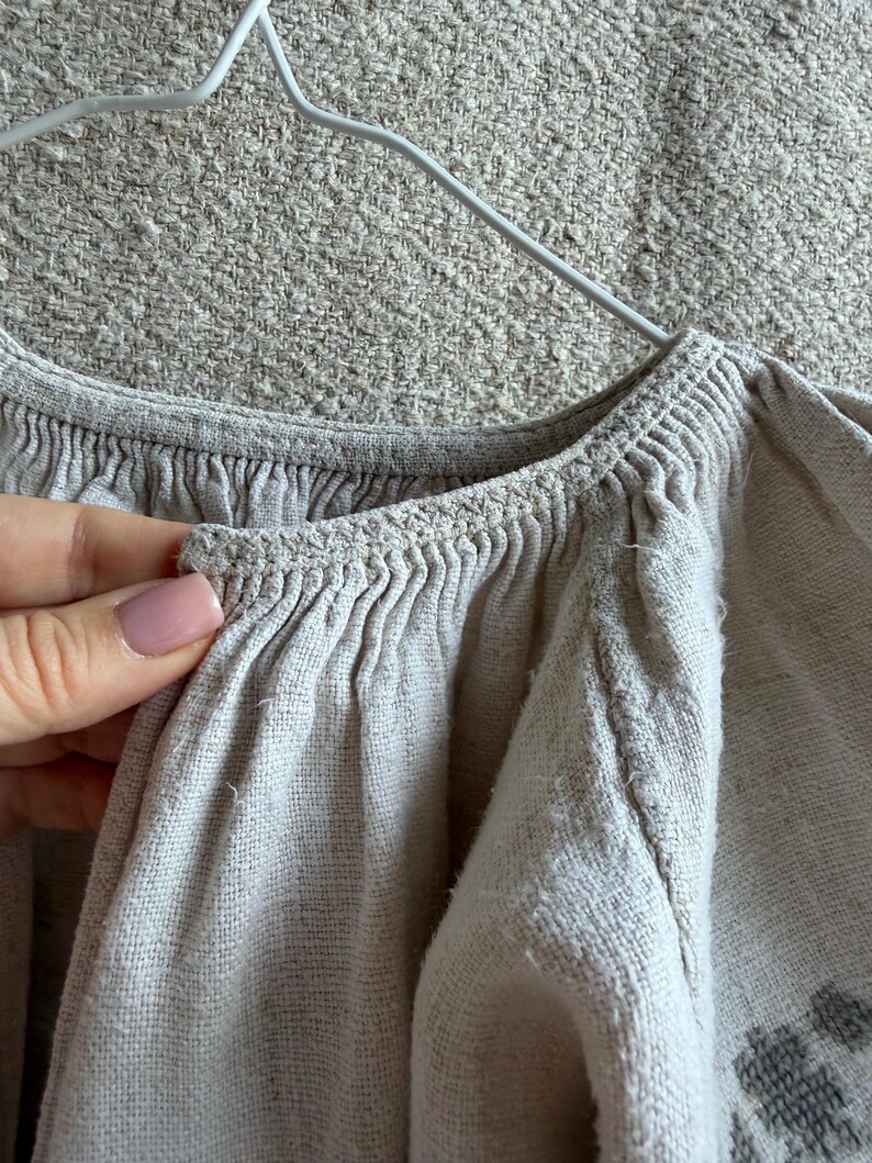 May include: A light beige linen blouse featuring a gathered neckline with intricate crochet detailing along the collar edge. The garment has long, voluminous sleeves and a textured fabric weave, displayed on a white wire hanger against a neutral background.