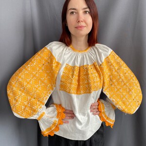 May include: A white blouse with long, puffy sleeves featuring intricate yellow embroidered patterns. The sleeves have a crocheted trim.
