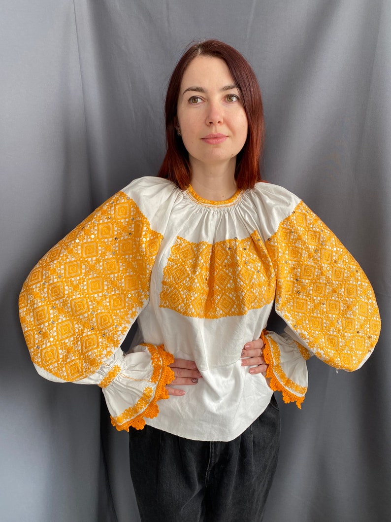 May include: A white blouse with long, puffy sleeves featuring intricate yellow embroidered patterns. The sleeves have a crocheted trim.