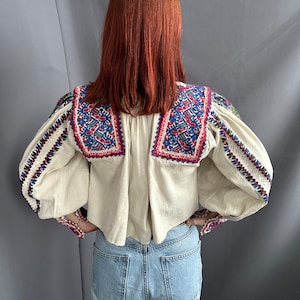 May include: A white peasant blouse with embroidered details in red, blue, and green. The blouse has long sleeves with a gathered cuff and a slightly cropped hem.