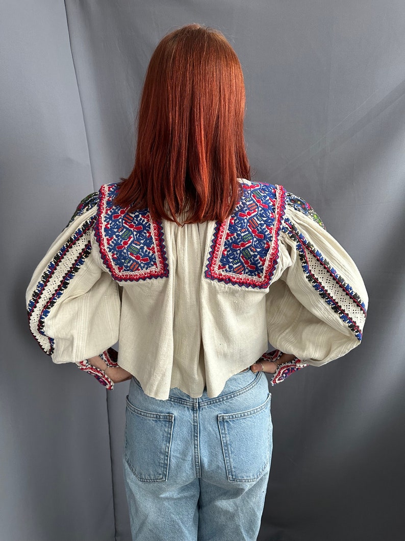 May include: A white peasant blouse with embroidered details in red, blue, and green. The blouse has long sleeves with a gathered cuff and a slightly cropped hem.