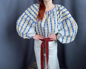 May include: A white embroidered blouse with blue and yellow stripes and a long white skirt with a red and yellow woven belt.