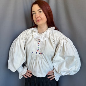 Op de afbeelding: Een wit boerenblouse met gepofte mouwen en een gerimpelde kraag. De blouse heeft kleurrijke geborduurde details en knopen.