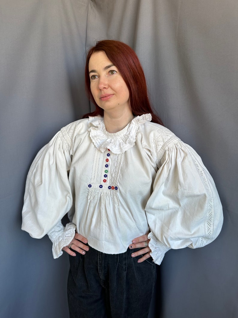 Op de afbeelding: Een wit boerenblouse met gepofte mouwen en een gerimpelde kraag. De blouse heeft kleurrijke geborduurde details en knopen.