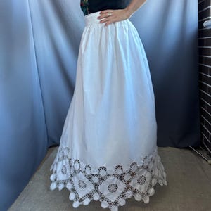 May include: A white skirt with a wide waistband and a full, flowing silhouette. The hemline features a detailed, openwork lace design. The skirt is made of a lightweight fabric, suitable for warm weather.