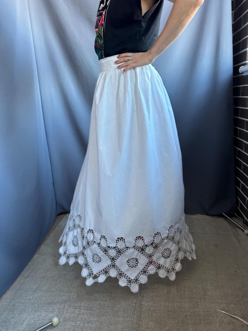 May include: A white skirt with a wide waistband and a full, flowing silhouette. The hemline features a detailed, openwork lace design. The skirt is made of a lightweight fabric, suitable for warm weather.