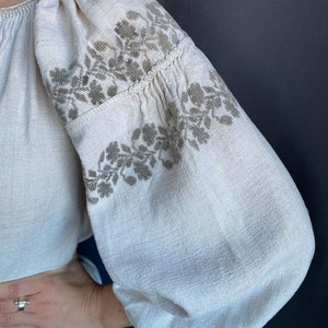 May include: A long-sleeved blouse made of off-white, textured linen fabric. The garment features voluminous, gathered sleeves adorned with intricate, muted olive-green floral embroidery patterns across the shoulders and upper arm sections.