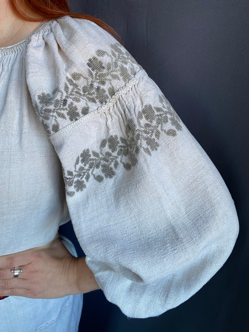 May include: A long-sleeved blouse made of off-white, textured linen fabric. The garment features voluminous, gathered sleeves adorned with intricate, muted olive-green floral embroidery patterns across the shoulders and upper arm sections.