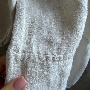 May include: A close-up view of a light beige, textured fabric garment sleeve. The material features a visible, coarse weave with a slightly fuzzy surface. A horizontal seam runs across the lower portion of the sleeve, secured with thick, visible stitching.