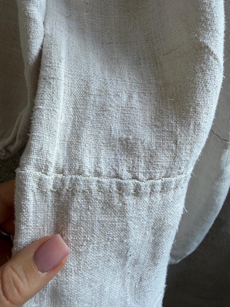 May include: A close-up view of a light beige, textured fabric garment sleeve. The material features a visible, coarse weave with a slightly fuzzy surface. A horizontal seam runs across the lower portion of the sleeve, secured with thick, visible stitching.