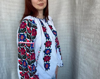 May include: White embroidered blouse with a floral pattern in red, blue, and green. The blouse has long sleeves and a round neckline.
