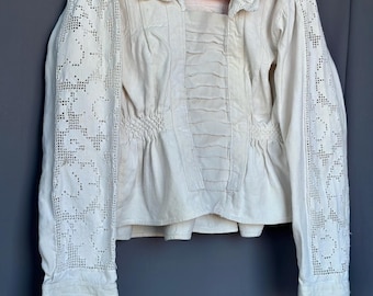 May include: An off-white long-sleeve blouse featuring intricate lace panels on the sleeves, a wide collar with scalloped edges, and a central panel with horizontal pleats. The waist is gathered with smocking, and the cuffs have lace trim.