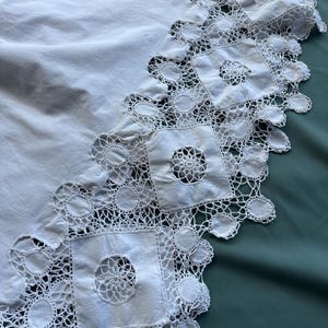 May include: A white, vintage-style tablecloth with a detailed, openwork lace border. The border features square and circular lace patterns, creating a delicate and ornate edge. The tablecloth is made of a soft, white fabric.