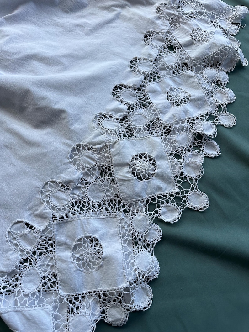 May include: A white, vintage-style tablecloth with a detailed, openwork lace border. The border features square and circular lace patterns, creating a delicate and ornate edge. The tablecloth is made of a soft, white fabric.