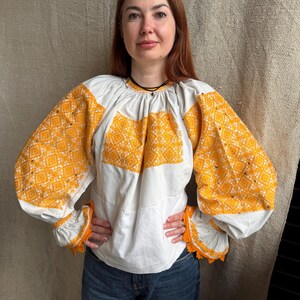 May include: A white long-sleeved blouse featuring intricate yellow geometric embroidery across the chest and voluminous sleeves. The garment has a gathered round neckline and cuffs finished with a decorative yellow crocheted lace trim.