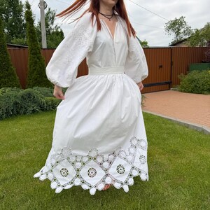 May include: A white dress with long, embroidered sleeves and a full skirt. The dress features a V-neckline and a decorative, openwork hem. The woman is standing on green grass.