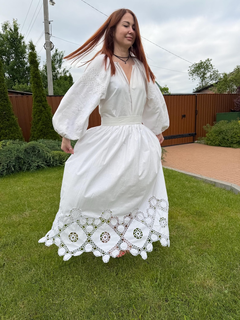 May include: A white dress with long, embroidered sleeves and a full skirt. The dress features a V-neckline and a decorative, openwork hem. The woman is standing on green grass.