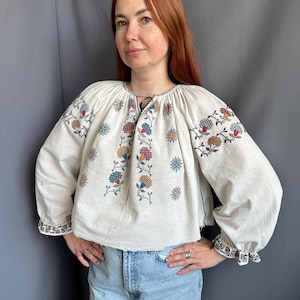 May include: A white linen blouse with embroidered floral designs in shades of red, blue, yellow, and green. The blouse has a gathered neckline and long sleeves with embroidered cuffs.