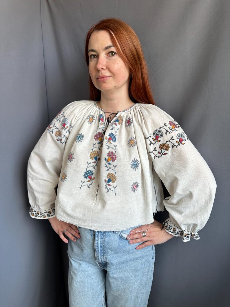 May include: A white linen blouse with embroidered floral designs in shades of red, blue, yellow, and green. The blouse has a gathered neckline and long sleeves with embroidered cuffs.