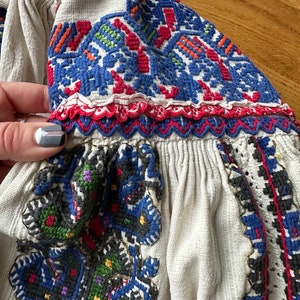May include: A white linen blouse with colorful embroidered details. The embroidery features geometric patterns in shades of blue, red, green, and yellow.